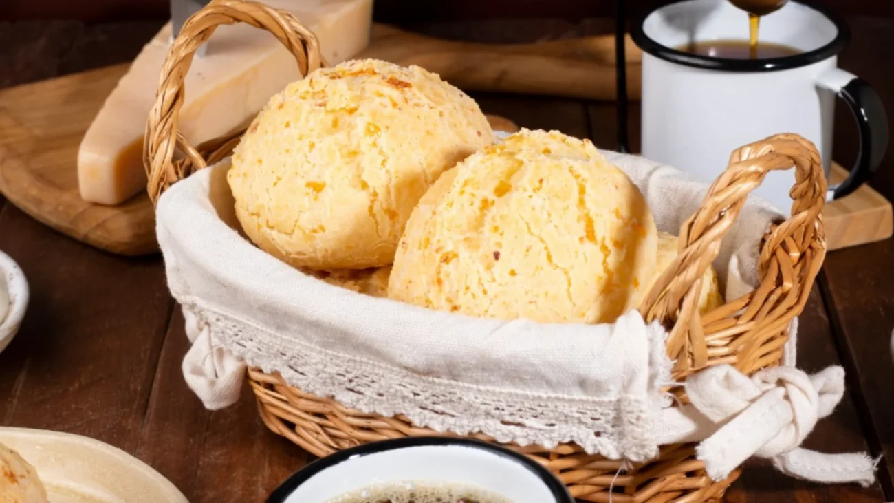 Como comer pão de queijo de formas criativas: pães de queijo dourados em cestinha com café e queijo ao fundo sobre mesa de madeira